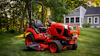 Red zero-turn radius riding mower on manicured lawn with residential house and trees in background