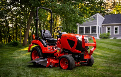 Red zero-turn radius riding mower on manicured lawn with residential house and trees in background