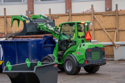 Green telehandler lifting waste materials into blue dumpster at construction site