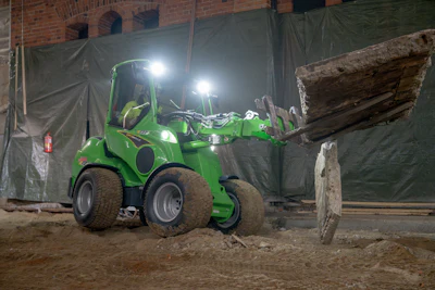 Green skid steer loader lifting wooden beam at illuminated construction site at night