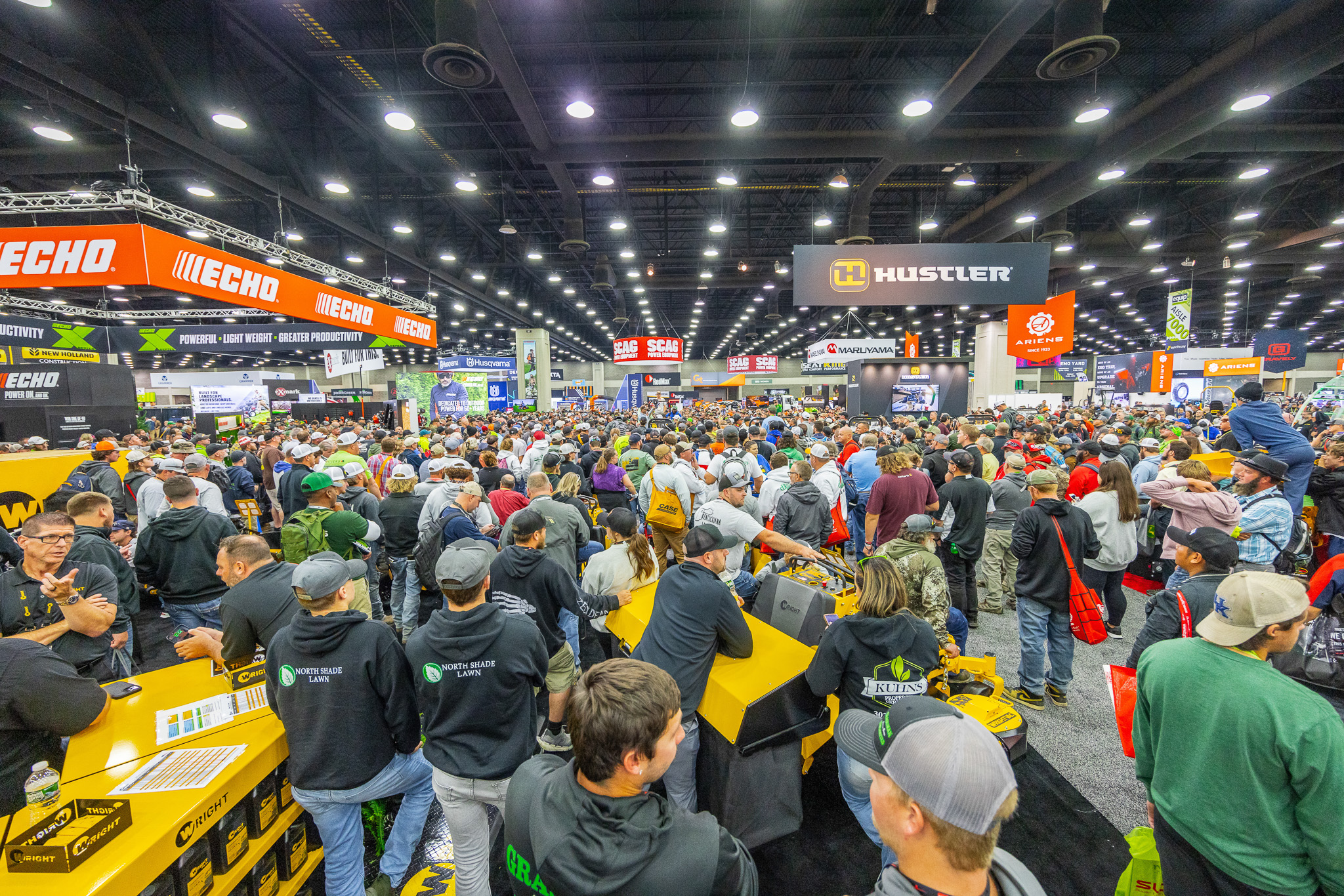 Crowded outdoor power equipment trade show with yellow machinery, orange and black brand signage, and hundreds of attendees in a large industrial venue.