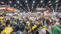 Crowded outdoor power equipment trade show with yellow machinery, orange and black brand signage, and hundreds of attendees in a large industrial venue.
