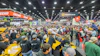 Crowded outdoor power equipment trade show with yellow machinery, orange and black brand signage, and hundreds of attendees in a large industrial venue.