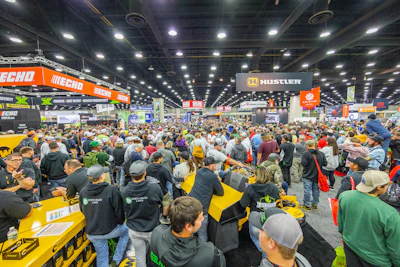 Crowded outdoor power equipment trade show with yellow machinery, orange and black brand signage, and hundreds of attendees in a large industrial venue.