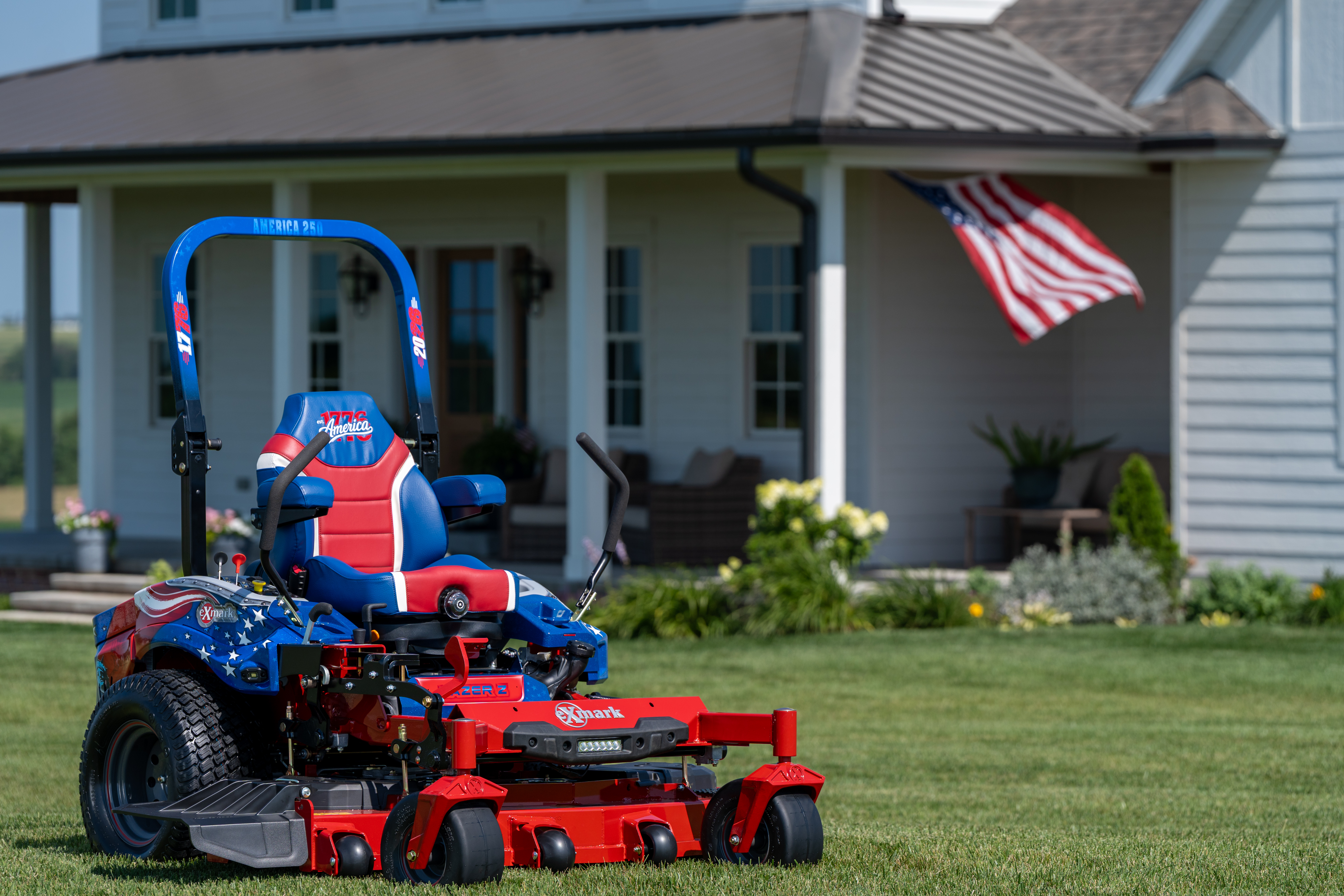 Patriotic red and blue zero-turn radius mower on residential lawn with American flag and house in background