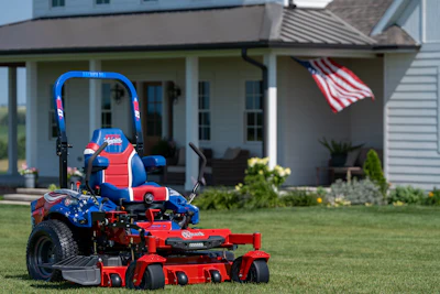 Patriotic red and blue zero-turn radius mower on residential lawn with American flag and house in background