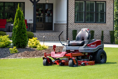 Red and gray zero-turn radius riding mower on manicured lawn in front of brick home with landscaping