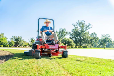 Man operating red Exmark zero-turn mower on manicured lawn with trees and water in background