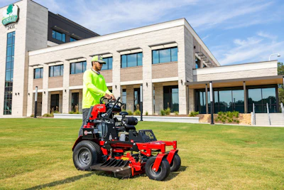 Grounds keeper operating red zero-turn mower on lawn in front of modern institutional building