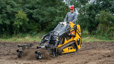 Operator using yellow Caterpillar skid steer loader to till soil on construction site surrounded by trees