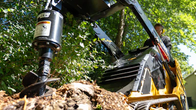 Yellow wood chipping machine grinding tree branches with operator in background surrounded by green foliage