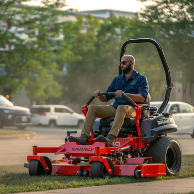 Man operating red Gravely zero-turn mower on residential street with trees and parked cars in background