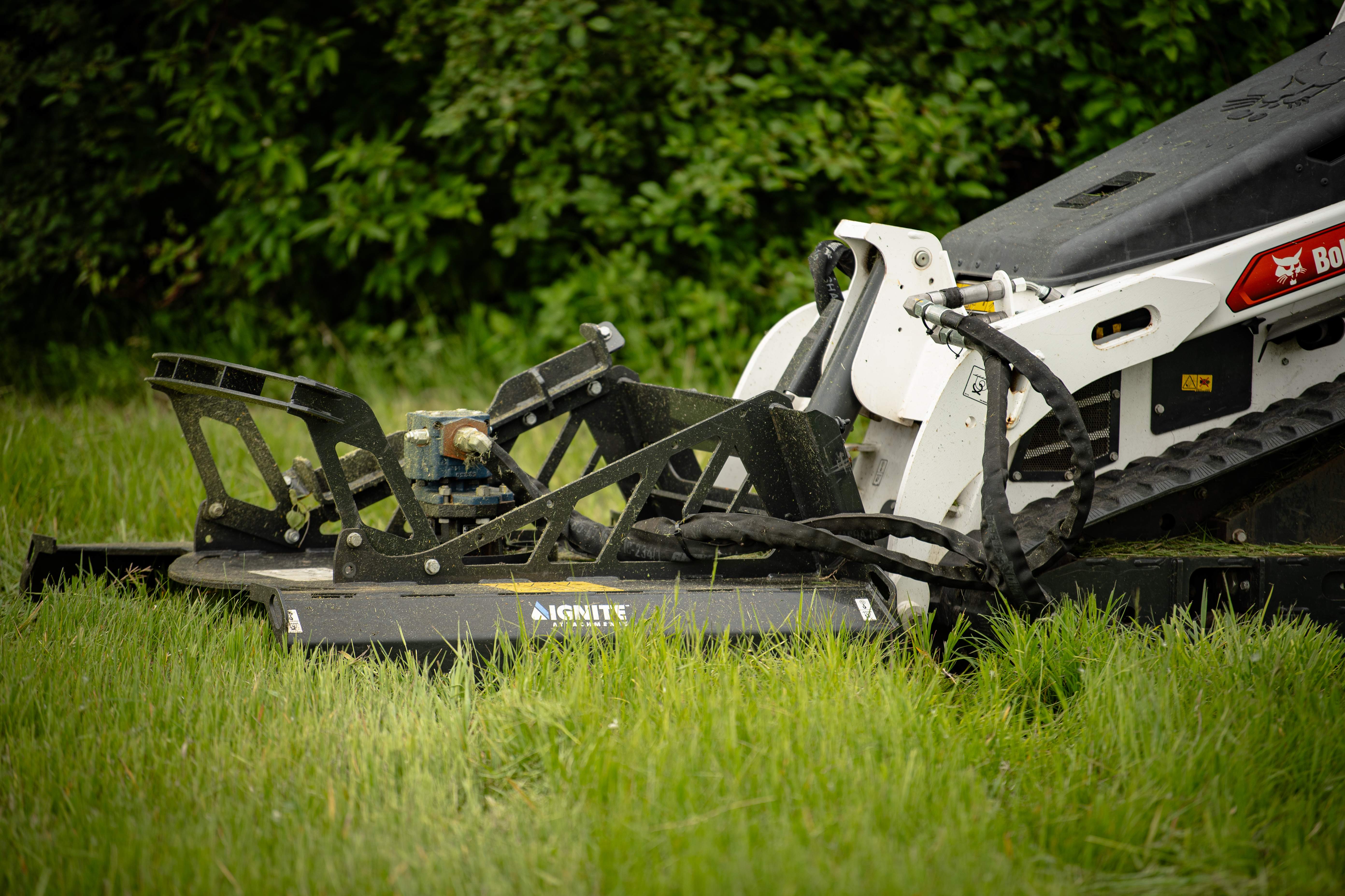 Bobcat skid steer loader with rotary mower attachment cutting grass in a lawn with green foliage background