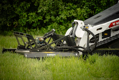 Bobcat skid steer loader with rotary mower attachment cutting grass in a lawn with green foliage background