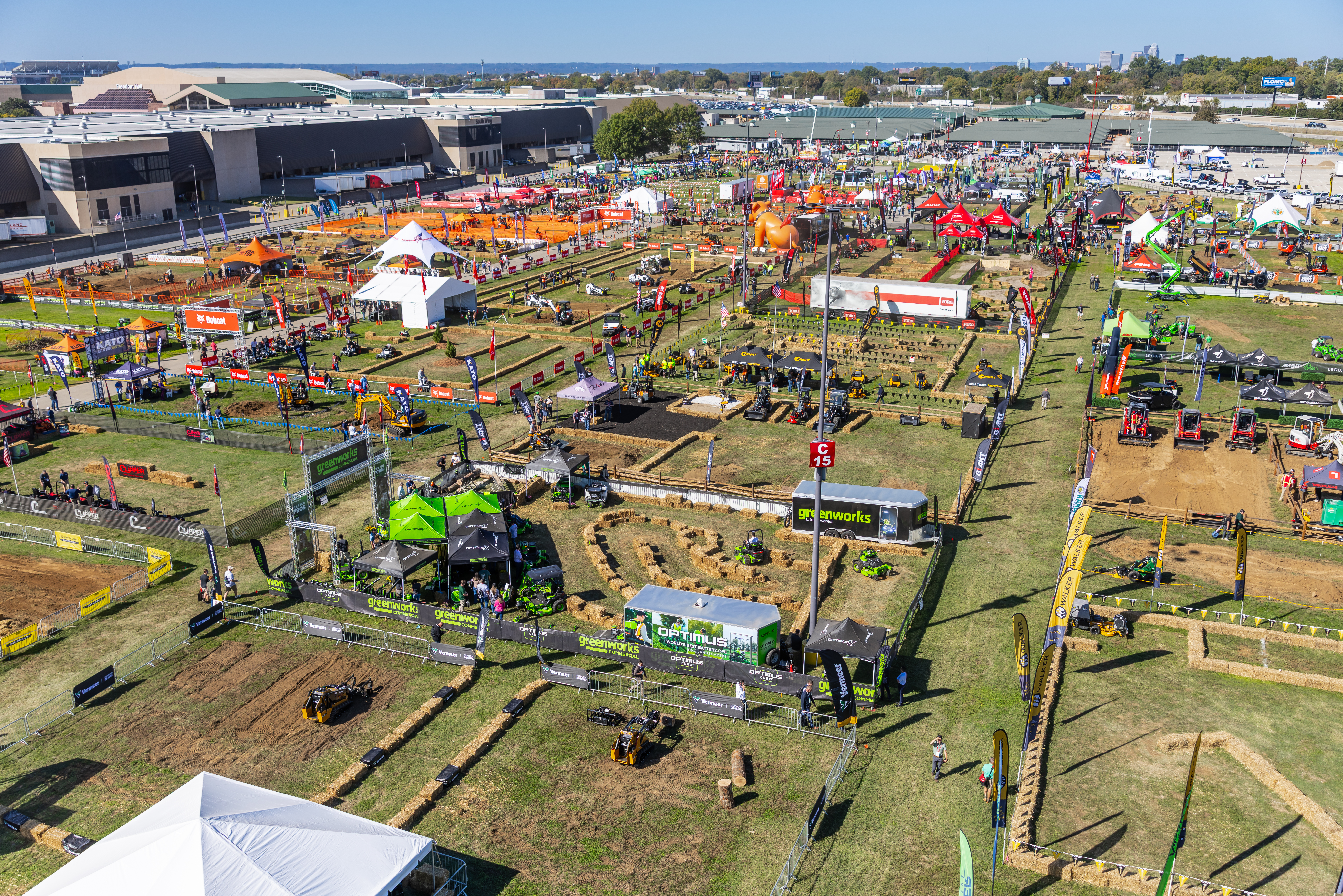 Aerial view of large agricultural equipment exhibition with machinery displays, vendor tents, and crowds at outdoor fairground.