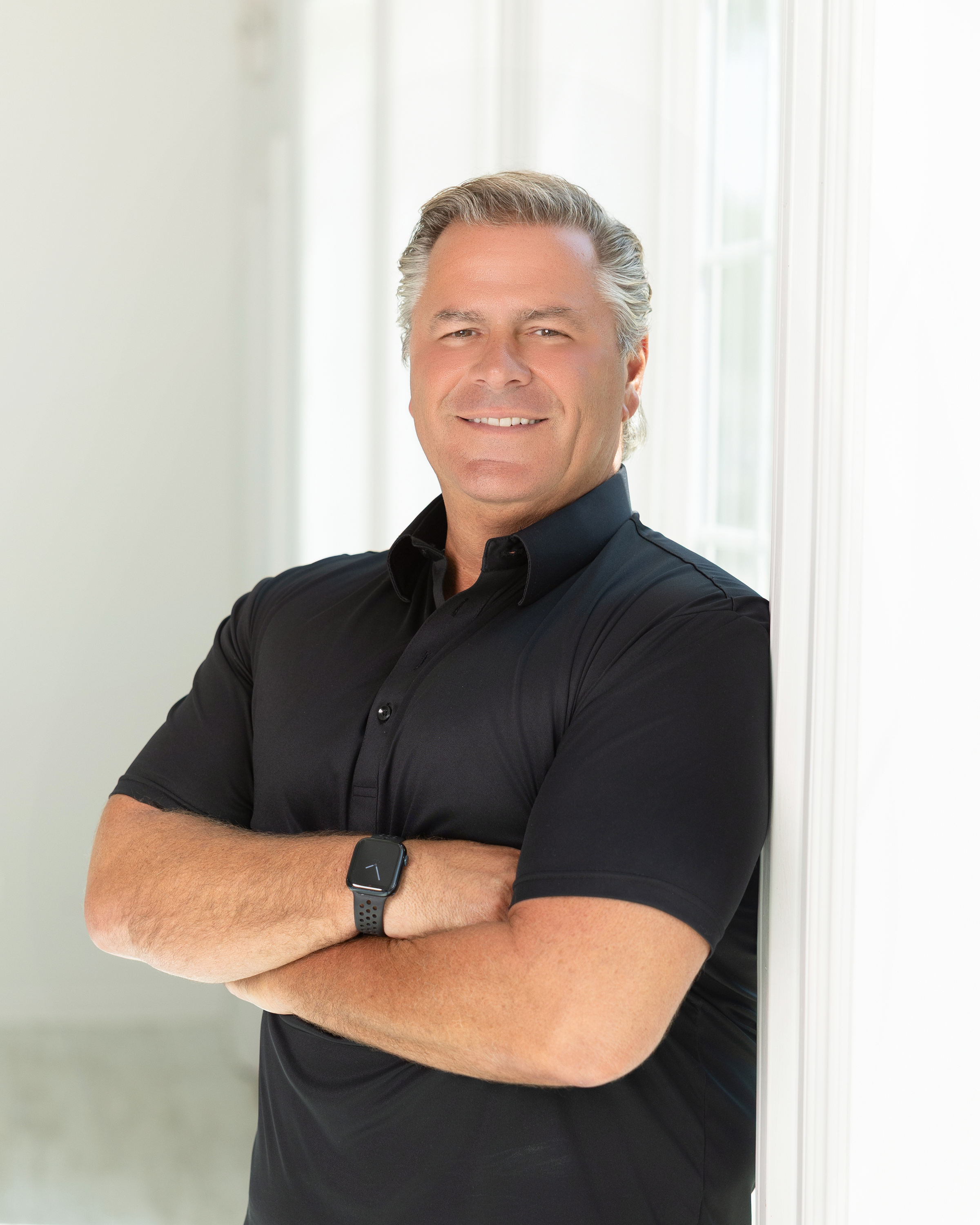 Professional headshot of a man in black polo shirt with arms crossed, smiling against white background