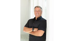 Professional headshot of a man in black polo shirt with arms crossed, smiling against white background