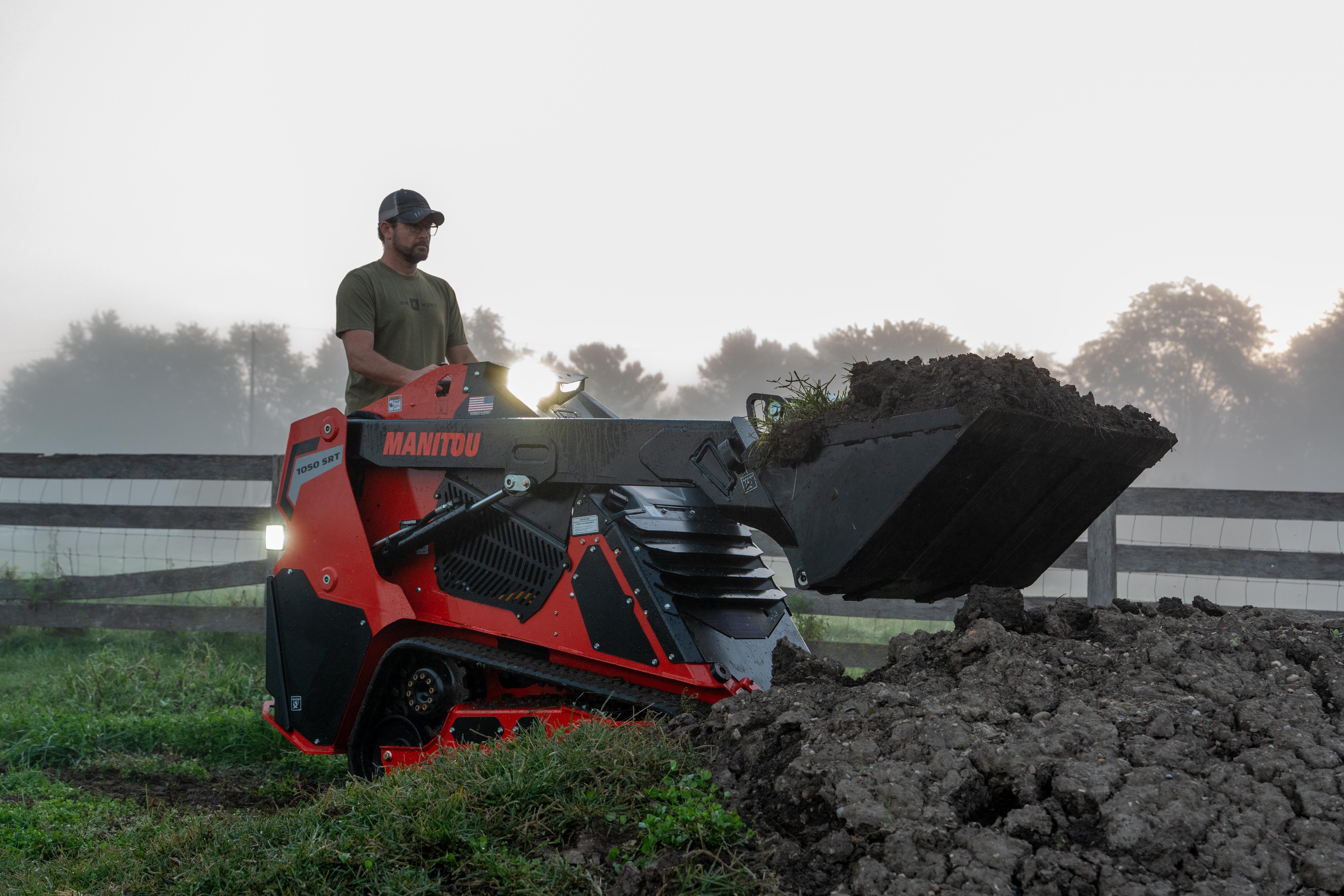 Man operating red Manitou telehandler loading dark soil into bucket on misty farm field