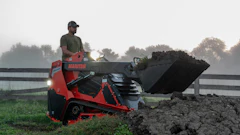 Man operating red Manitou telehandler loading dark soil into bucket on misty farm field