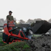 Man operating red Manitou telehandler loading dark soil into bucket on misty farm field