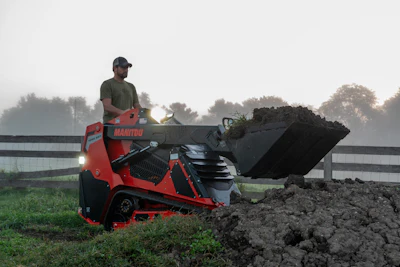 Man operating red Manitou telehandler loading dark soil into bucket on misty farm field