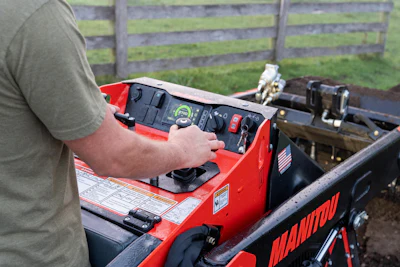Person operating red Manitou telehandler control panel in farm setting with wooden fence background