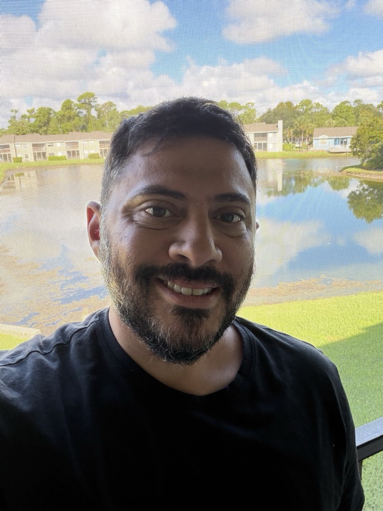 Man with beard smiling at camera with waterfront buildings and green lawn in background