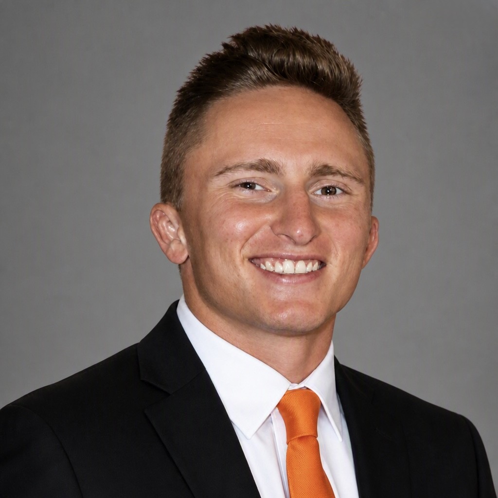 Professional headshot of smiling man in black suit and orange tie against gray background Headshot