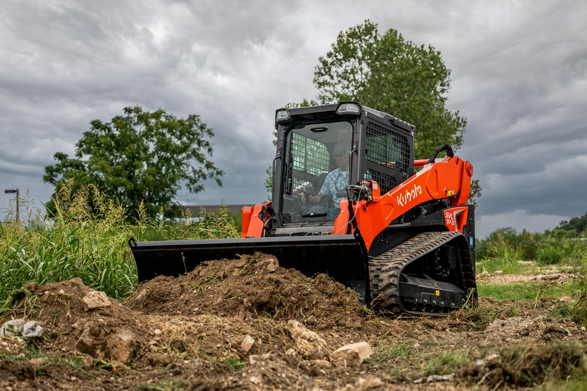 Orange Kubota compact track loader with blade pushing soil pile in rural field under cloudy sky