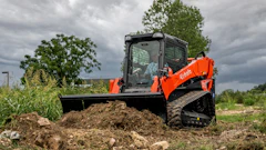 Orange Kubota compact track loader with blade pushing soil pile in rural field under cloudy sky