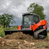 Orange Kubota compact track loader with blade pushing soil pile in rural field under cloudy sky