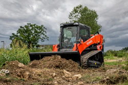 Orange Kubota compact track loader with blade pushing soil pile in rural field under cloudy sky