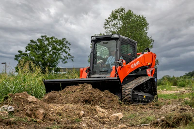 Orange Kubota compact track loader with blade pushing soil pile in rural field under cloudy sky
