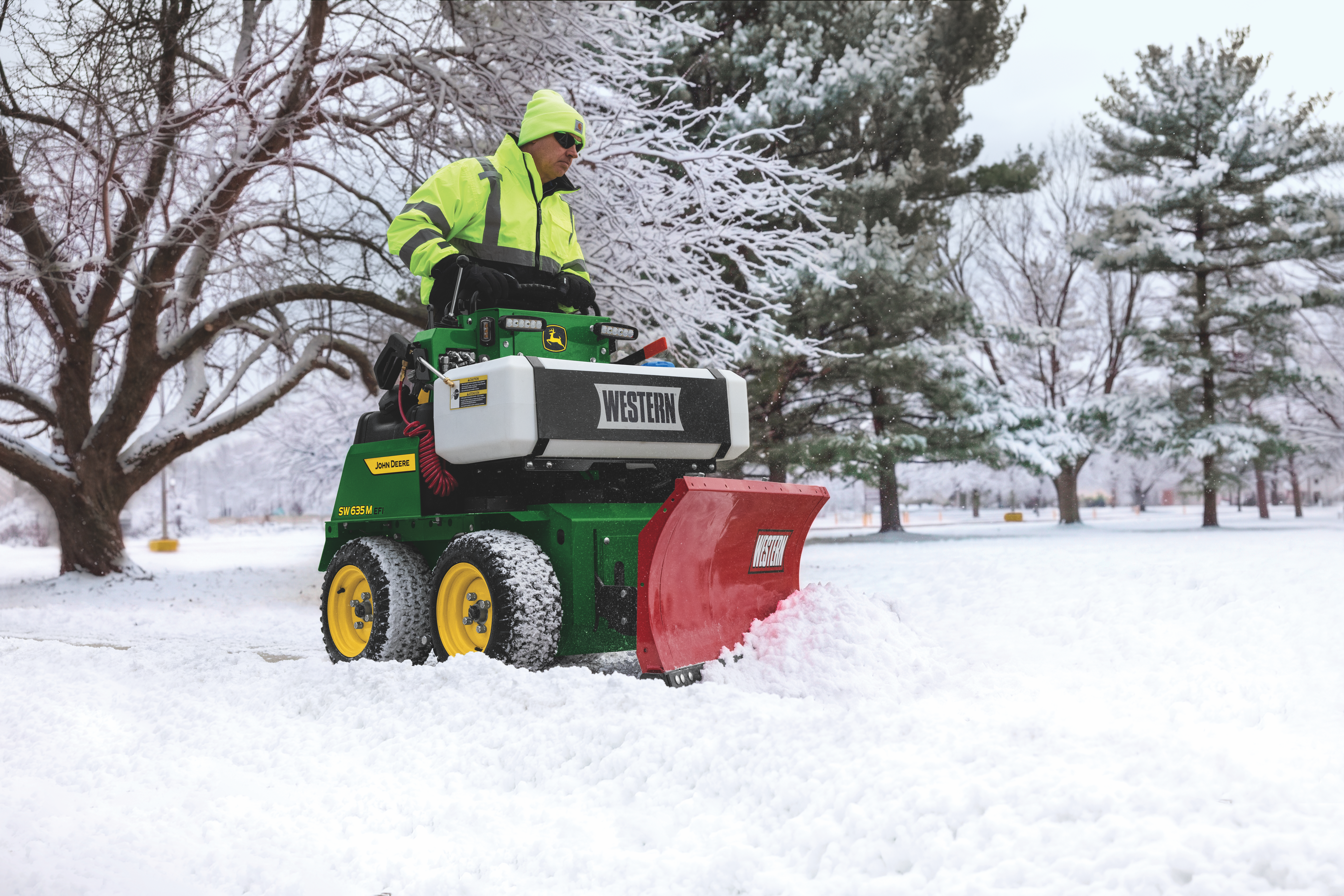 Worker in safety gear operating a green John Deere snow plow with red blade clearing snow from a winter park path