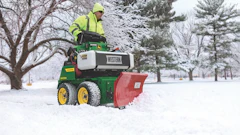 Worker in safety gear operating a green John Deere snow plow with red blade clearing snow from a winter park path