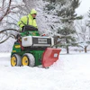 Worker in safety gear operating a green John Deere snow plow with red blade clearing snow from a winter park path