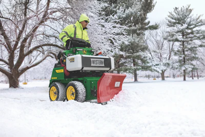 Worker in safety gear operating a green John Deere snow plow with red blade clearing snow from a winter park path