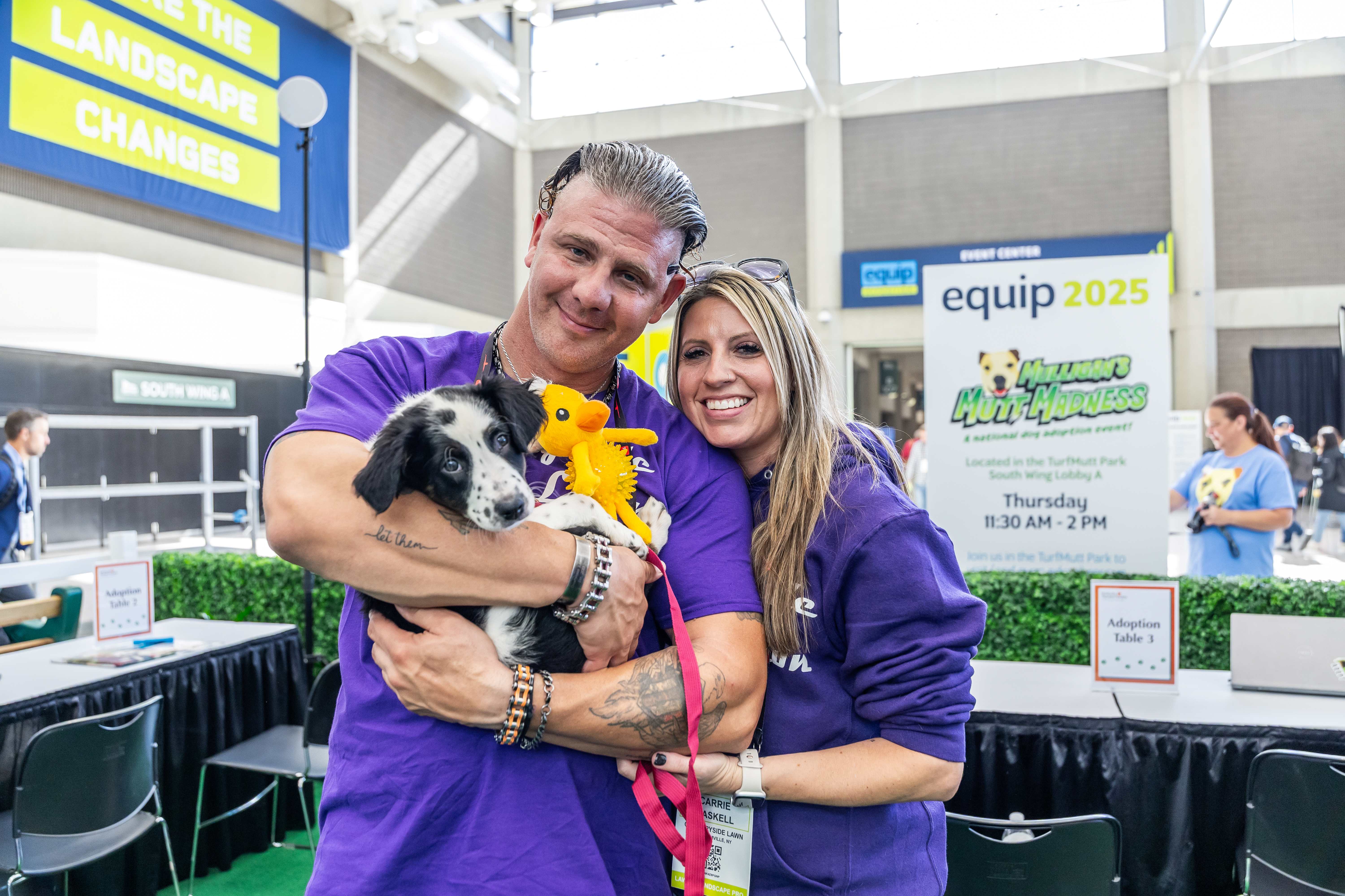 Two people in purple shirts hold a black and white dog at an indoor equip 2025 event