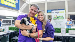 Two people in purple shirts hold a black and white dog at an indoor equip 2025 event