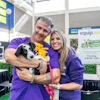 Two people in purple shirts hold a black and white dog at an indoor equip 2025 event