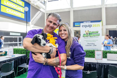 Two people in purple shirts hold a black and white dog at an indoor equip 2025 event